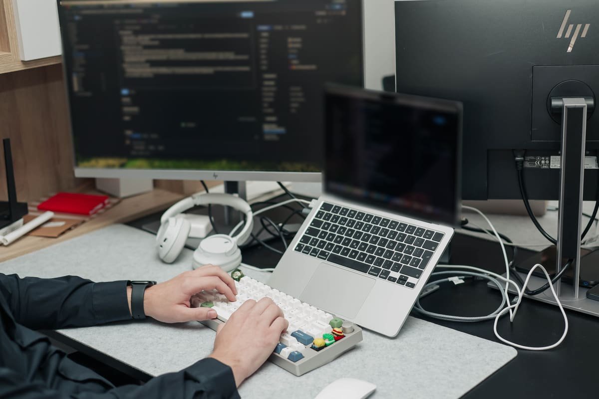 Hände auf einer mechanischen Tastatur vor einem Laptop und einem Monitor mit Code